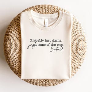 Puede incluir: Camiseta color crema con el texto negro "Probably just gonna jingle some of the way. I'm tired.". La camiseta est&aacute; doblada y colocada sobre una superficie circular tejida.