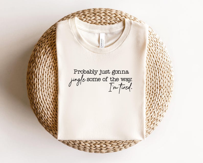 Puede incluir: Camiseta color crema con el texto negro "Probably just gonna jingle some of the way. I'm tired.". La camiseta est&aacute; doblada y colocada sobre una superficie circular tejida.