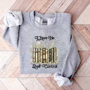 May include: A heather gray sweatshirt with the text "I Have No Shelf Control" in black script. The graphic features a stack of vintage-style books surrounded by floral elements. The sweatshirt has long sleeves.