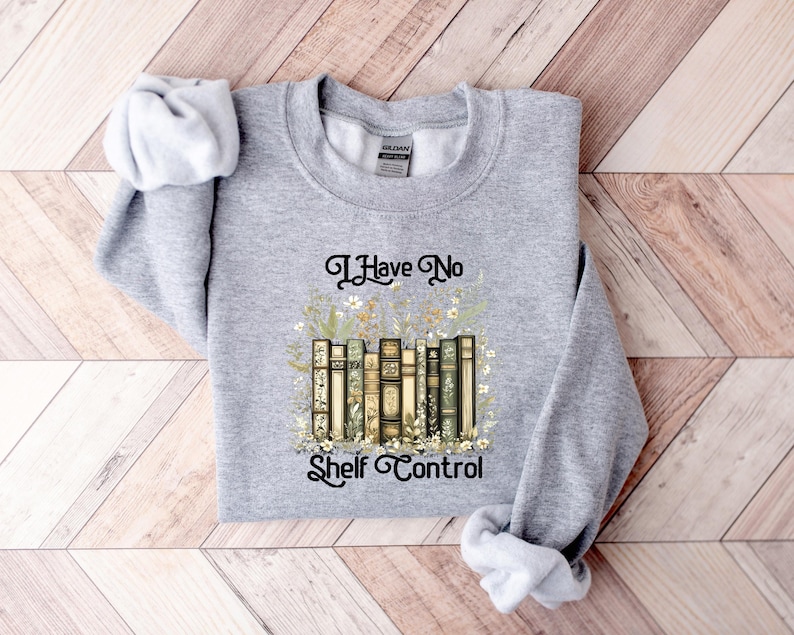 May include: A heather gray sweatshirt with the text "I Have No Shelf Control" in black script. The graphic features a stack of vintage-style books surrounded by floral elements. The sweatshirt has long sleeves.