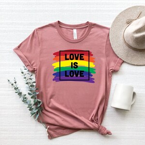 May include: A dusty rose t-shirt with the text "LOVE IS LOVE" in a black box over a rainbow-colored brushstroke design. A neutral-toned hat and a white mug are visible in the background. The shirt is folded at the bottom.
