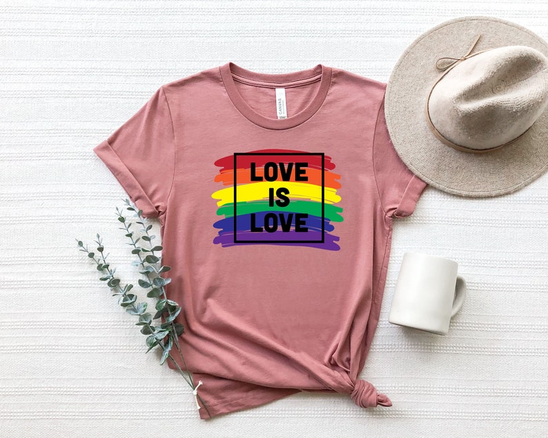 May include: A dusty rose t-shirt with the text "LOVE IS LOVE" in a black box over a rainbow-colored brushstroke design. A neutral-toned hat and a white mug are visible in the background. The shirt is folded at the bottom.
