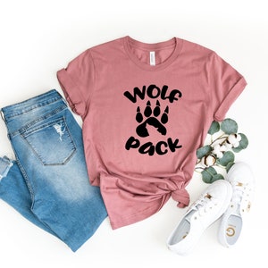 May include: A mauve t-shirt with a black paw print and the text "Wolf Pack". The t-shirt is paired with blue jeans and white sneakers.