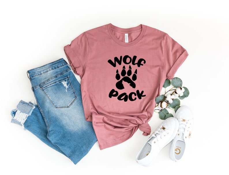 May include: A mauve t-shirt with a black paw print and the text "Wolf Pack". The t-shirt is paired with blue jeans and white sneakers.