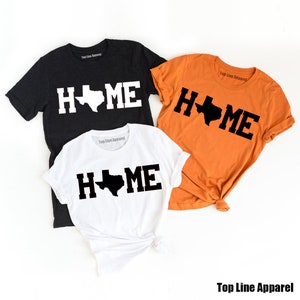 May include: Three t-shirts with the word "HOME" in black letters with a black outline of the state of Texas in the middle of the word. The shirts are black, white, and orange.
