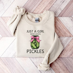 Puede incluir: Una sudadera beige con el texto "Just a girl who loves pickles" y un gr&aacute;fico de un frasco de pepinillos con un lazo rosa.