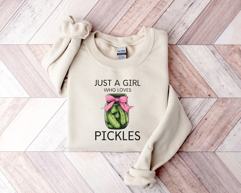 Puede incluir: Una sudadera beige con el texto "Just a girl who loves pickles" y un gr&aacute;fico de un frasco de pepinillos con un lazo rosa.
