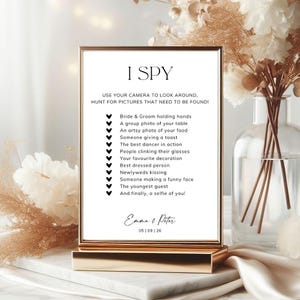 May include: A rectangular wedding I SPY game sign in a gold frame. The white card features black text listing photo tasks, ending with the names Emma & Peter and the date 05 | 09 | 26. The list includes items like bride and groom holding hands.