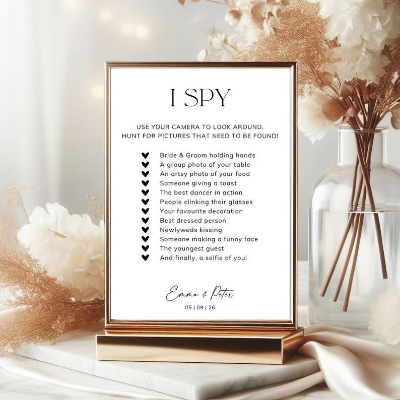 May include: A rectangular wedding I SPY game sign in a gold frame. The white card features black text listing photo tasks, ending with the names Emma & Peter and the date 05 | 09 | 26. The list includes items like bride and groom holding hands.