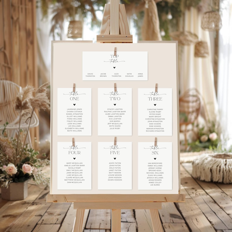 May include: A wooden easel displaying a seating chart for a wedding or event. The chart features a top card and six table cards, all with names in elegant black script. The background is a neutral beige, with decorative elements.
