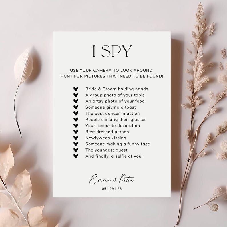May include: A white rectangular wedding I Spy game card with black text and heart icons. The card lists photo prompts for guests and features the names Emma & Peter with the date 05 | 09 | 26 at the bottom in a clean, black serif font.