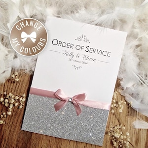 May include: A white wedding Order of Service card. The card features the text "ORDER OF SERVICE", the names "Kelly & Steven", and the date "26th March 2024". It has a silver glitter base, a pink ribbon, and a pink bow with a crystal.