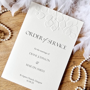 May include: White wedding order of service booklet with an embossed floral design. The text reads "ORDER of SERVICE" for the marriage of "DIANE JOHNSON & MARTIN SMITH" St Agnes Church, Glasgow 16.08.24.