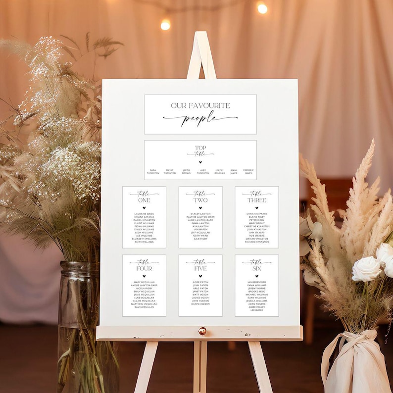 May include: A white wedding seating chart on an easel. The chart features the text "Our Favourite People" and "Top Table", with guest names listed under each table number. The design is minimalist with black text on a white background.
