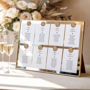 May include: A gold-framed wedding seating chart with eight white cards, each labeled with a table number and names. The cards are held in place by gold wax seals. Two champagne flutes are in the foreground.