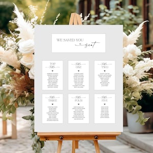 May include: A white seating chart on an easel with the text "WE SAVED YOU a seat". The chart is divided into sections labeled "Top Table" through "Table Five", each listing names. The background features floral arrangements.