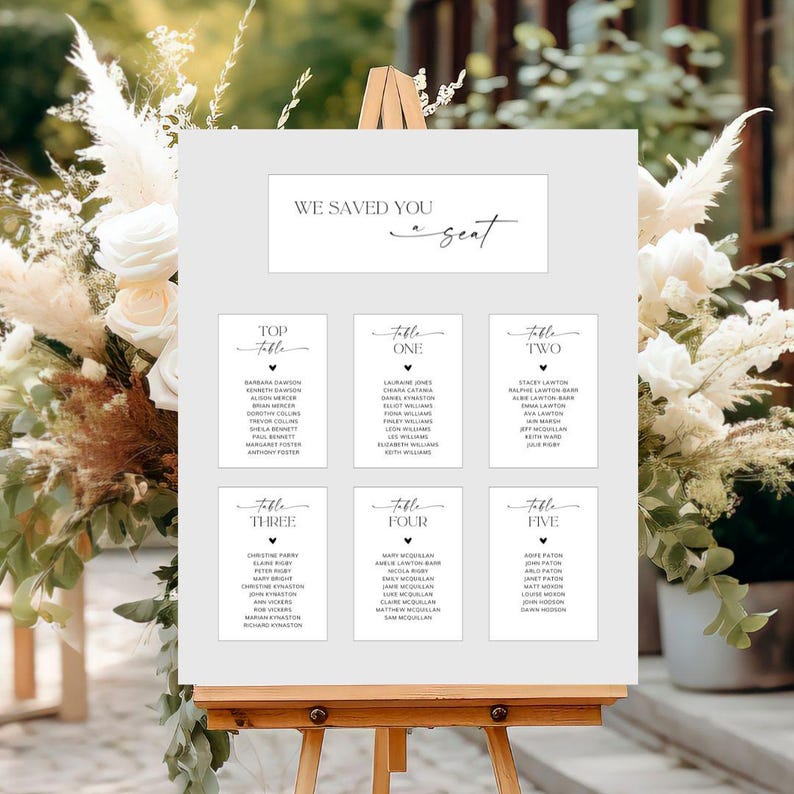 May include: A white seating chart on an easel with the text "WE SAVED YOU a seat". The chart is divided into sections labeled "Top Table" through "Table Five", each listing names. The background features floral arrangements.