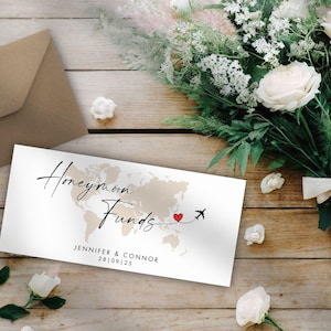 May include: A white card with the words "Honeymoon Funds" and a world map design. A red heart and airplane graphic are included. The names Jennifer & Connor and the date 28|09|25 are printed below. A brown envelope and a bouquet of white flowers are in the background.