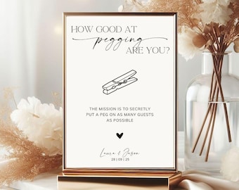 Puede incluir: Un letrero rectangular en un marco dorado con fondo blanco y texto negro que dice HOW GOOD AT pegging ARE YOU? con un dibujo lineal de una pinza de ropa, instrucciones para colocar secretamente una pinza a los invitados, un corazón y los nombres Laura & Jason.