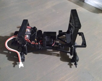May include: A black plastic chassis for a remote control car with a motor, wires, and a circuit board. The chassis is designed for off-road use and has a suspension system.