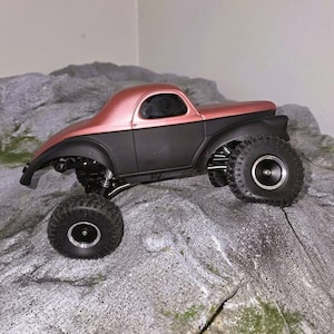 Pu&ograve; includere: Un veicolo radiocomandato di tipo crawler con carrozzeria superiore in oro rosa metallizzato e telaio inferiore nero opaco. Il veicolo &egrave; dotato di grandi pneumatici neri tassellati, cerchi argentati e componenti delle sospensioni visibili su una roccia.