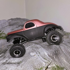 Pu&ograve; includere: Un veicolo radiocomandato di tipo crawler con carrozzeria superiore in oro rosa metallizzato e telaio inferiore nero opaco. Il veicolo &egrave; dotato di grandi pneumatici fuoristrada testurizzati con cerchi argentati e sospensioni a vista.