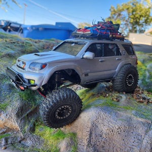 Könnte beinhalten: Ein silbernes ferngesteuertes Offroad-SUV-Modell mit großen schwarzen Stollenreifen, einem Frontschutzbügel und einem Dachträger, der mit roten Spanngurten befestigt ist. Das Fahrzeug verfügt über ein detailliertes Äußeres mit sichtbaren Federungselementen.
