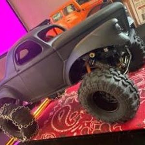 Pu&ograve; includere: Un modello di camion radiocomandato nero opaco con grandi pneumatici fuoristrada robusti. Lo pneumatico anteriore &egrave; dotato di catene da neve in metallo. Il veicolo presenta un sistema di sospensioni dettagliato con molle visibili e telaio scuro.