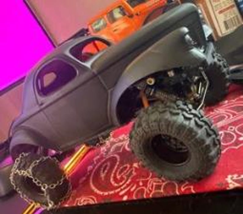 Pu&ograve; includere: Un modello di camion radiocomandato nero opaco con grandi pneumatici fuoristrada robusti. Lo pneumatico anteriore &egrave; dotato di catene da neve in metallo. Il veicolo presenta un sistema di sospensioni dettagliato con molle visibili e telaio scuro.