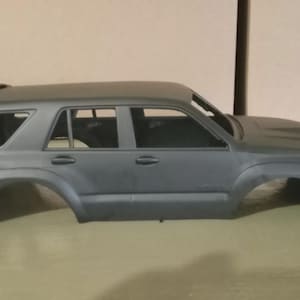 Może przedstawiać: Szary plastikowy model samochodu sportowo-użytkowego (SUV) z bagażnikiem dachowym i oknami. Modelowi brakuje kół i innych elementów. Pojazd ma około 30 cm długości.