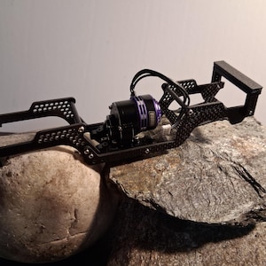 May include: Black RC car chassis with a purple and black motor. The chassis is made of metal and has a complex, open design with multiple mounting points. The motor has the word "NIORA" printed on it. The chassis is resting on a rock.