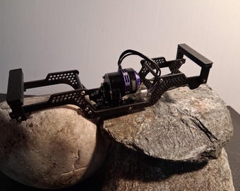 May include: Black RC car chassis with a purple and black motor. The chassis is made of metal and has a complex, open design with multiple mounting points. The motor has the word "NIORA" printed on it. The chassis is resting on a rock.