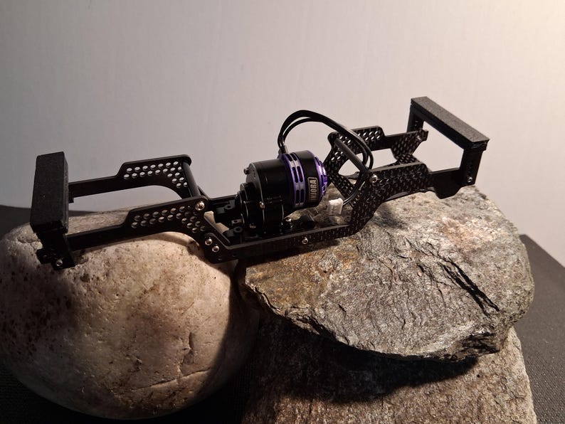May include: Black RC car chassis with a purple and black motor. The chassis is made of metal and has a complex, open design with multiple mounting points. The motor has the word "NIORA" printed on it. The chassis is resting on a rock.