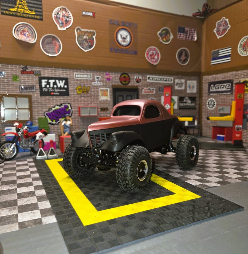 Pu&ograve; includere: Un modellino di camion personalizzato bicolore marrone e nero opaco, dotato di pneumatici fuoristrada sovradimensionati con cerchi argentati, una griglia anteriore e una cabina arrotondata, esposto in un diorama da garage.