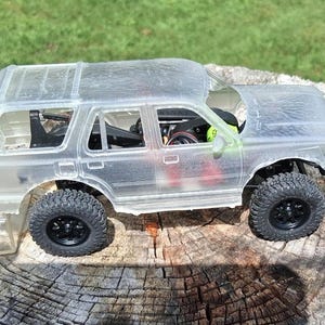 Könnte beinhalten: Eine klare, unlackierte Kunststoff-Karosserie für ein SUV-Modell mit detailliertem Innenraum, schwarzen Rädern mit strukturierten Gummireifen und einer sichtbaren Chassis-Struktur. Das Modell ist für ferngesteuerte Fahrzeuge konzipiert und zeigt interne mechanische Komponenten.