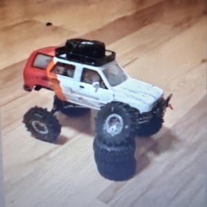 Könnte beinhalten: Ein weiß-rotes Geländewagen-Spielzeug mit einem schwarzen Dachträger, der ein Ersatzrad trägt. Das Fahrzeug hat große, grobstollige schwarze Reifen, wobei ein Vorderrad auf einem Stapel aus zwei weiteren schwarzen Reifen ruht.