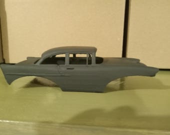 Può includere: Una carrozzeria per modellino d'auto in grigio opaco non verniciato, che mostra il profilo laterale distintivo di un veicolo classico degli anni '50. Il modello presenta passaruota aperti, una linea del tetto con finestrini e un lungo cofano.