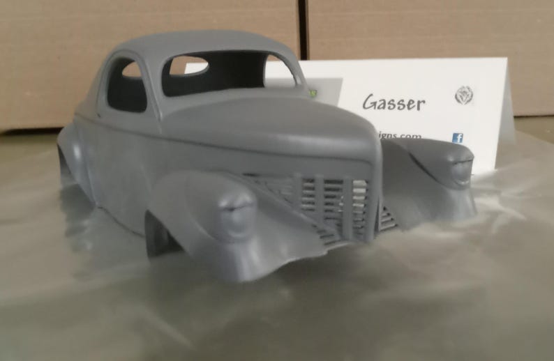 Pu&ograve; includere: Una carrozzeria per modellino d'auto grigia e non verniciata, caratterizzata da una classica silhouette coup&eacute; con parafanghi anteriori prominenti, una griglia verticale e fari arrotondati. Dietro il modello c'&egrave; un cartoncino con la scritta Gasser.