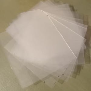 以下が含まれることがあります： 透明でクリアなプラスチック製のスリーブの束で、個々のシートを表示するために扇形に広げられています。スリーブは正方形で、書類や写真を保管するために設計されているようです。背景は明るい緑色です。