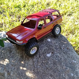 Könnte beinhalten: Ein dunkelrotes SUV-Modellauto mit glänzender Oberfläche, schwarzen Stoßstangen, silberfarbenen Felgen und grobstolligen Geländereifen. Das Fahrzeug verfügt über ein offenes Kabinendesign ohne Fenster, einen Dachträger und seitliche Außenspiegel.