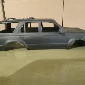 Könnte beinhalten: Eine matte dunkelgraue Kunststoff-Karosserie eines SUV-Modellautos. Das Äußere verfügt über einen Dachgepäckträger, Seitenspiegel, Türgriffe und offene Fensterrahmen. Die Radkästen sind leer und die Motorhaube zeigt ein dezentes Reliefdetail.
