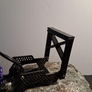 May include: A black, angular, 3D-printed model, possibly a component for a small vehicle or machine. The model has a rectangular top and a cross-braced support structure. A purple and black motor with the text "INJORA" is visible.