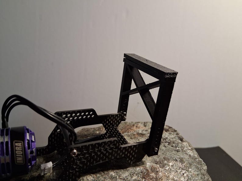 May include: A black, angular, 3D-printed model, possibly a component for a small vehicle or machine. The model has a rectangular top and a cross-braced support structure. A purple and black motor with the text "INJORA" is visible.