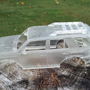 Könnte beinhalten: Eine klare, durchscheinende Modellauto-Karosserie aus Kunststoff mit der detaillierten Silhouette eines SUV, inklusive Dachgepäckträger, Seitenfenstern und Türverkleidungen. Das unlackierte, halbtransparente Material zeigt die Linien.