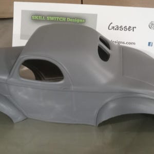 Pu&ograve; includere: Una carrozzeria per modellino d'auto grigia non verniciata con un design classico in stile gasser, caratterizzata da un tetto arrotondato, parafanghi curvi e finestrini aperti. Dietro il modello c'&egrave; un cartoncino bianco con la scritta SKILL SWITCH Designs e Gasser.