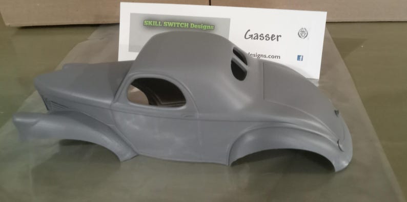 Pu&ograve; includere: Una carrozzeria per modellino d'auto grigia non verniciata con un design classico in stile gasser, caratterizzata da un tetto arrotondato, parafanghi curvi e finestrini aperti. Dietro il modello c'&egrave; un cartoncino bianco con la scritta SKILL SWITCH Designs e Gasser.