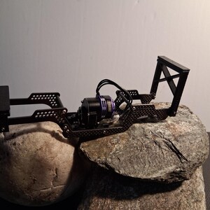 May include: A black, open-frame chassis for a small remote-controlled vehicle. The chassis has a motor with black wires and a purple casing. The frame is mounted on a gray rock, with a white background.