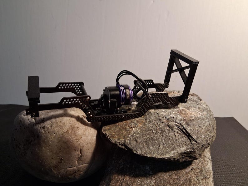 May include: A black, open-frame chassis for a small remote-controlled vehicle. The chassis has a motor with black wires and a purple casing. The frame is mounted on a gray rock, with a white background.