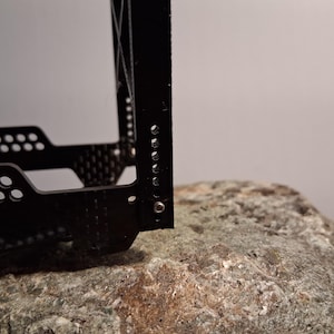 May include: Close-up of a black metal object with a perforated design and several small holes. The object is resting on a textured, gray rock. The background is a plain, light gray.