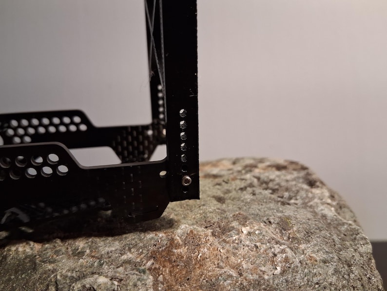 May include: Close-up of a black metal object with a perforated design and several small holes. The object is resting on a textured, gray rock. The background is a plain, light gray.